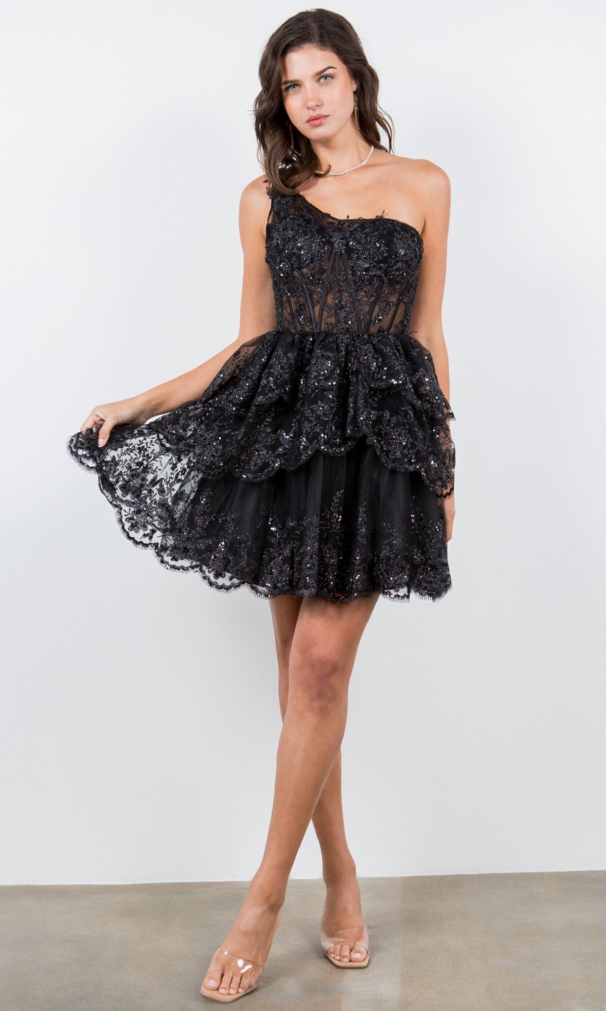 Woman wearing the One-Shoulder Short Tiered Homecoming Dress 5132J—black mini dress with a tiered skirt and sheer bodice—styled for semi-formal occasions, standing against a white background.